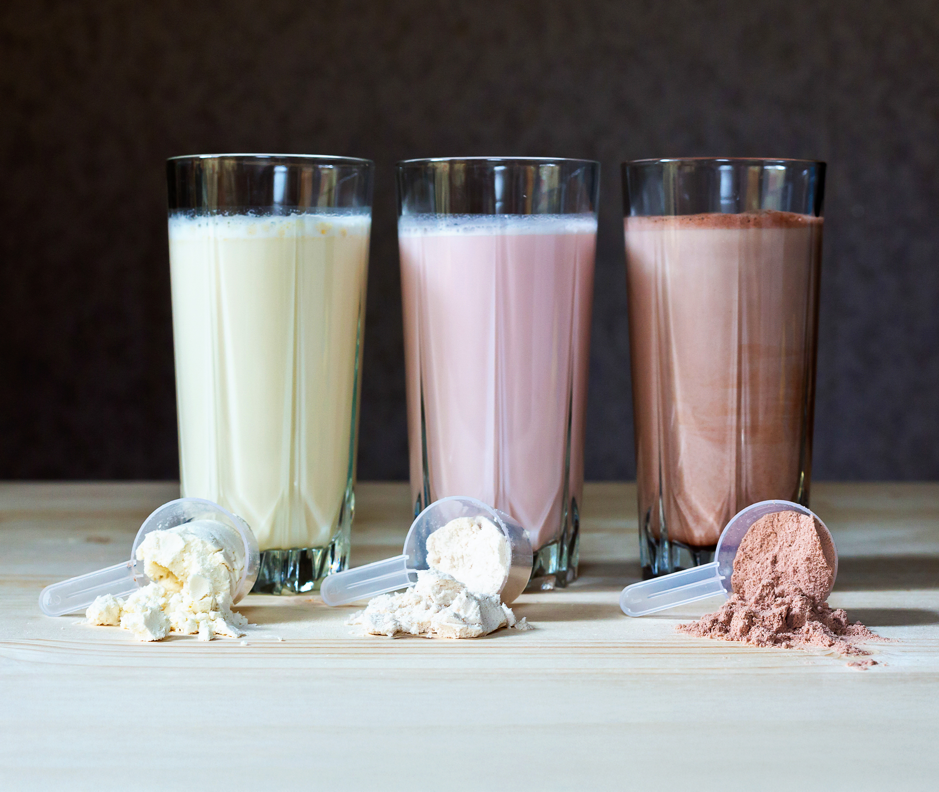 Wie viele Protein Shakes am Tag trinken für den Muskelaufbau Drei Gläser mit Proteinshakes und jeweils ein Scoop mit Pulver im Vordergrund